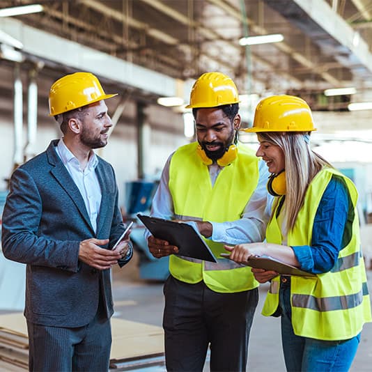 Three construction professionals in helmets and vests discuss a project in an industrial setting, reviewing documents and holding tablets.
