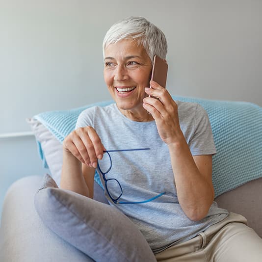 Woman with short grey hair, holding glasses, smiles while talking on a smartphone, sitting on a sofa with a light blue blanket.