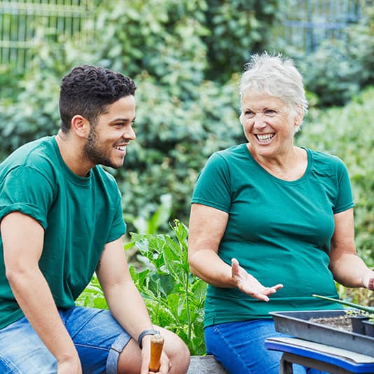 An older woman and a young man, both in green shirts, joyfully chat while gardening outdoors.