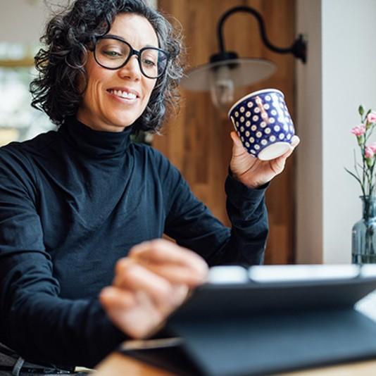 Woman working on tablet device holding a cup in kitchen