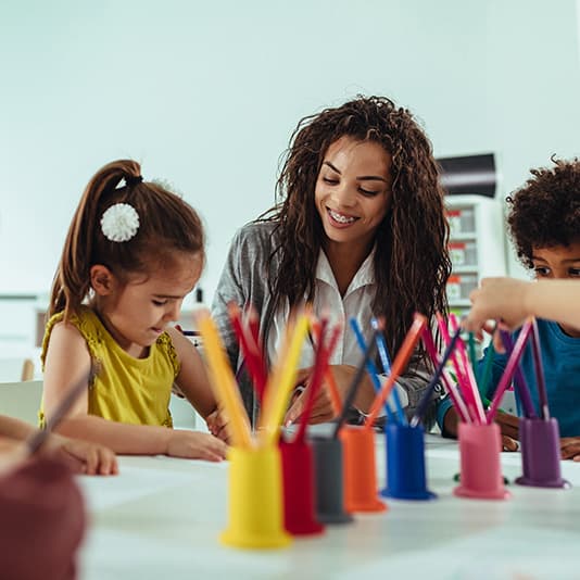 A woman smiles while helping children with a drawing activity at a table, surrounded by colourful pencil holders