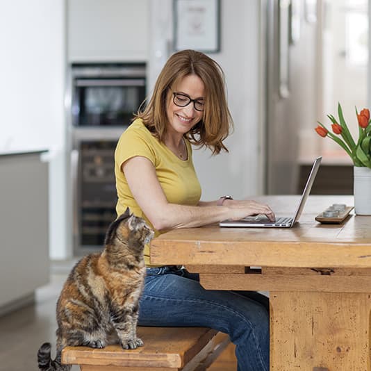 A woman in a yellow shirt works on a laptop at a wooden table, with a cat sitting nearby and a vase of tulips in the background.