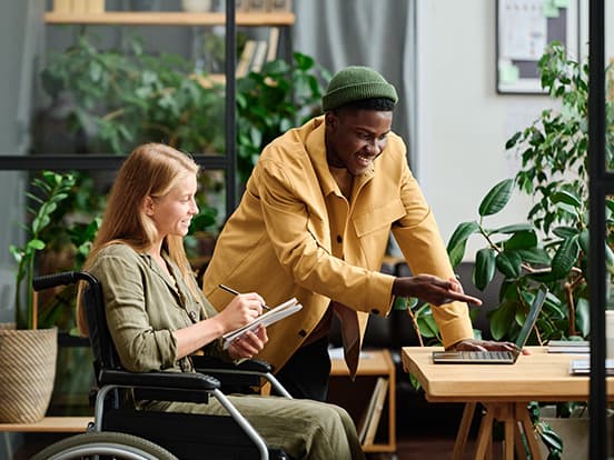 A woman in a wheelchair takes notes while a man in a yellow jacket points at a laptop. They are in a bright, plant-filled office space.