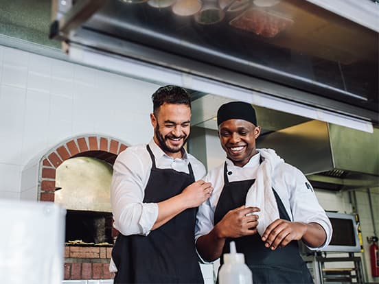 Two chefs in aprons share a laugh in a kitchen. One wears a headscarf and holds a towel. A pizza oven is visible in the background.
