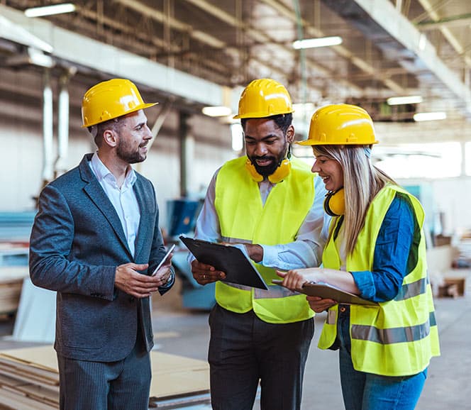 Three construction workers in hard hats and vests discuss plans on a clipboard inside a warehouse setting.