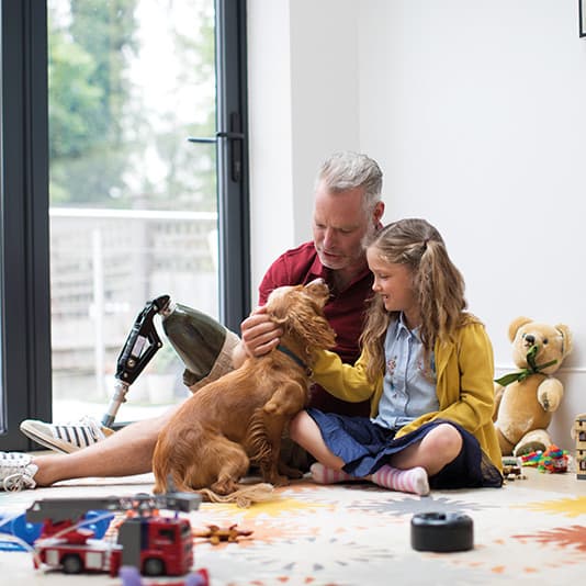 A man with a prosthetic leg and a young girl sit on the floor, petting a dog. Toys and a teddy bear are scattered around them