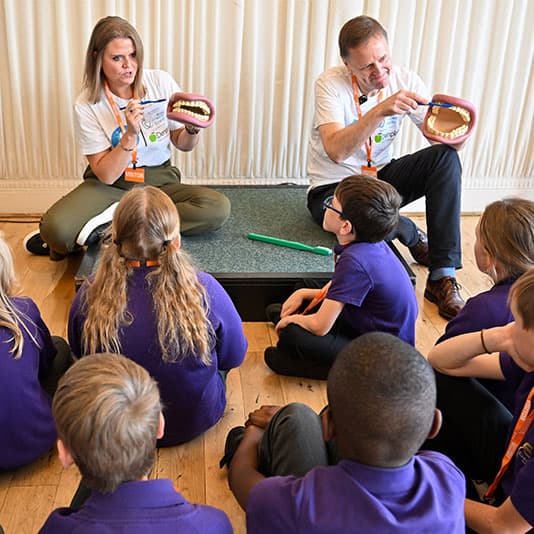 Two adults demonstrate dental care using large teeth models to a group of children sitting on the floor.