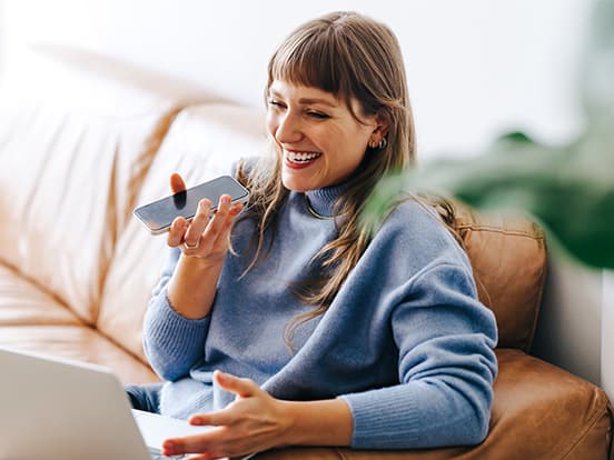 Woman in blue jumper, sitting on a sofa, smiling while using a smartphone and laptop