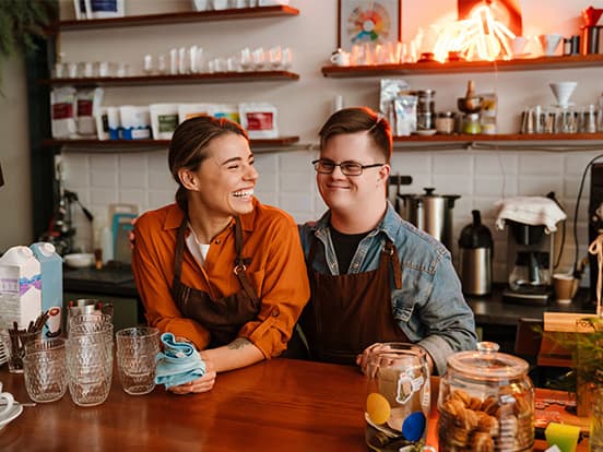 Two baristas in aprons, smiling and standing behind a counter in a cozy, well-stocked coffee shop.