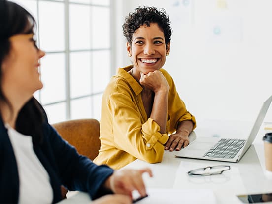 Two women smiling at a table, one in a yellow shirt near a laptop, bright room with a large window in the background.