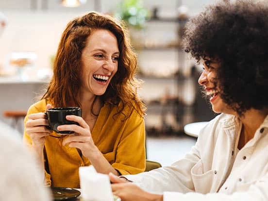 Two women laughing and chatting over coffee in a cozy café, one holding a black mug.