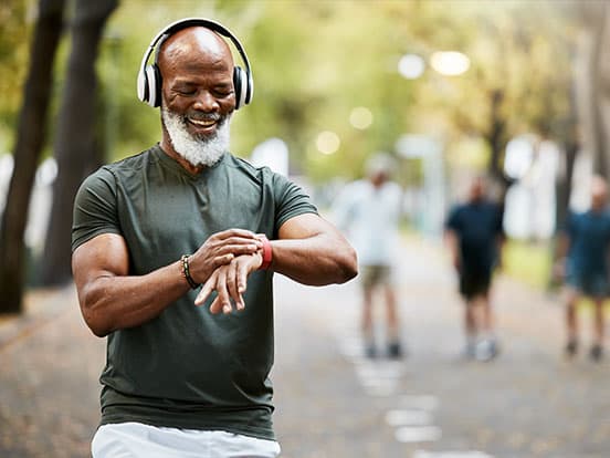Smiling older man with white beard wearing headphones, checking a smartwatch while walking on a tree-lined path.