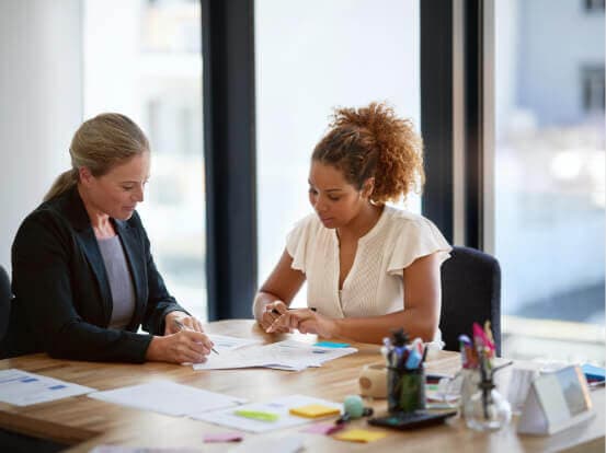 Two women sit at a desk reviewing documents, with office supplies and notes scattered around. A large window is in the background.