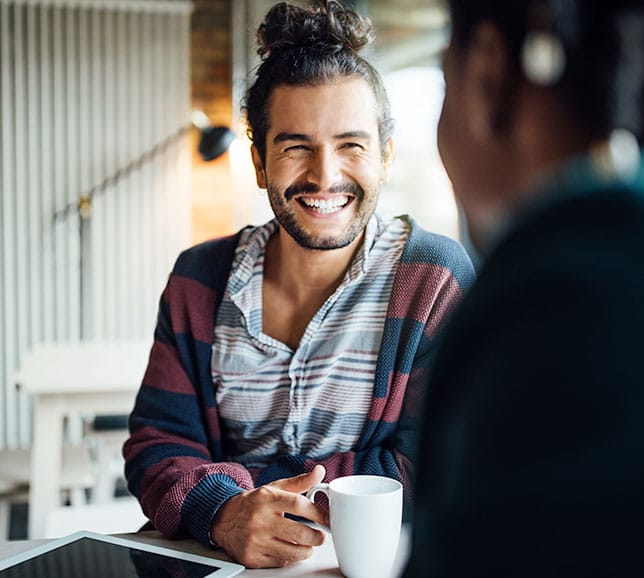 Man holding a mug and smiling at a friend across a table