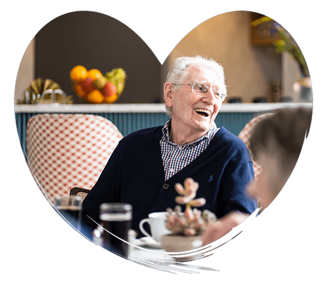 Elderly man smiling while sitting at a dining table with a coffee cup, plants, and fruit in the background.