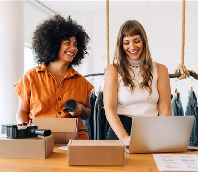 Two women working happily at a table, one using a laptop and the other holding a barcode scanner, with racks of clothes in the background.